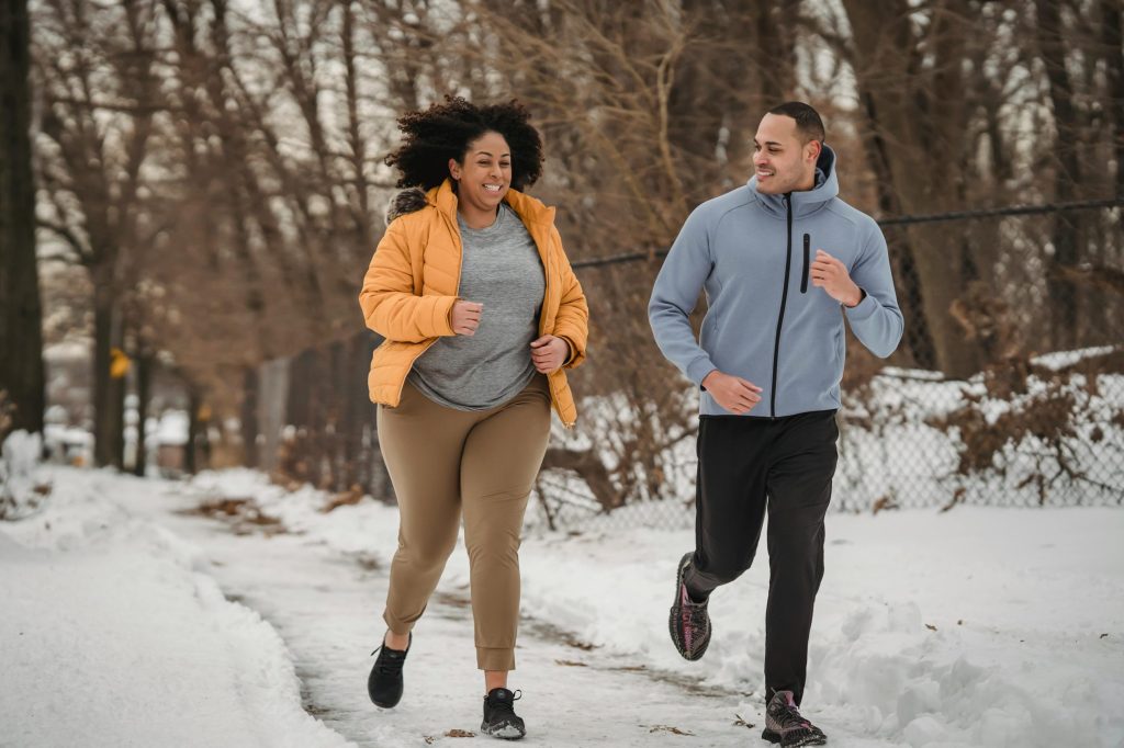 Full length smiling ethnic male fitness trainer and plus sized black female in warm jacket jogging together on snowy pathway in frozen winter park